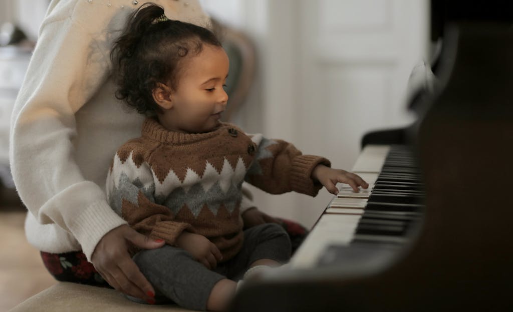 A cute toddler in a cozy sweater explores piano keys while sitting on a guardian's lap indoors.
