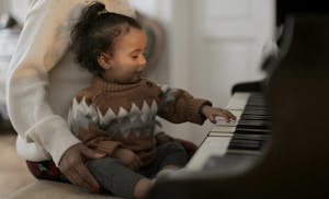 A cute toddler in a cozy sweater explores piano keys while sitting on a guardian's lap indoors.