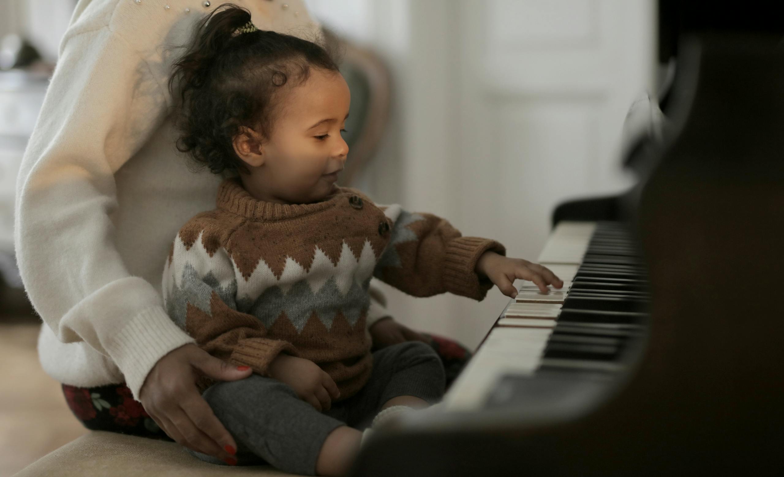 A cute toddler in a cozy sweater explores piano keys while sitting on a guardian's lap indoors.
